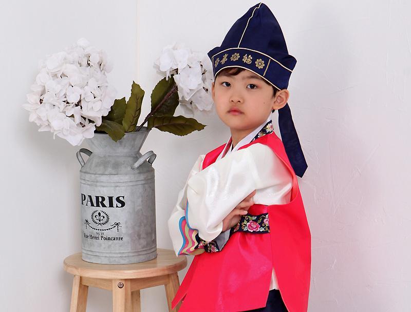 Young boy wearing a bright pink hanbok and navy hat posing next to flowers