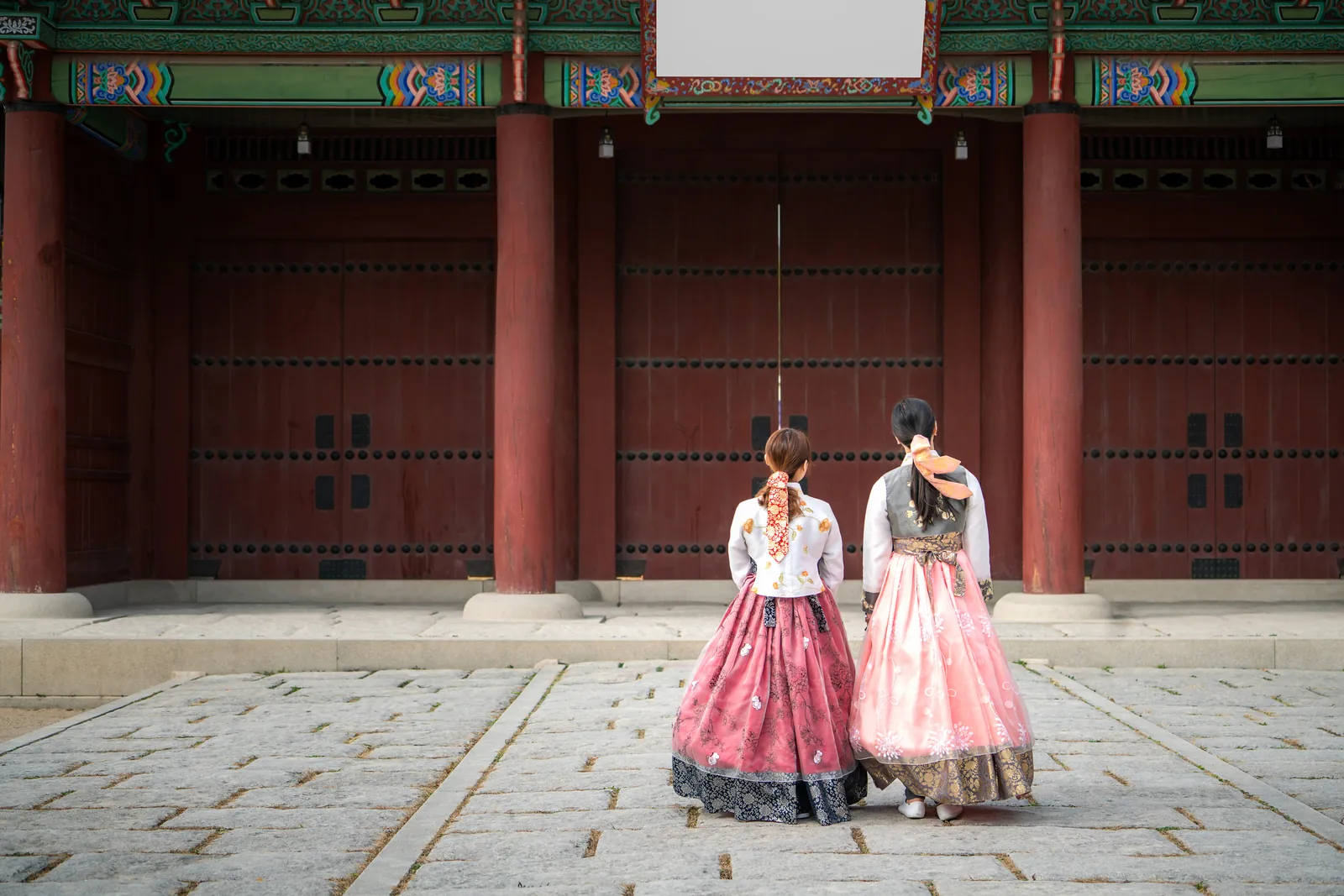 Two women in hanbok standing before a tall red wooden gate